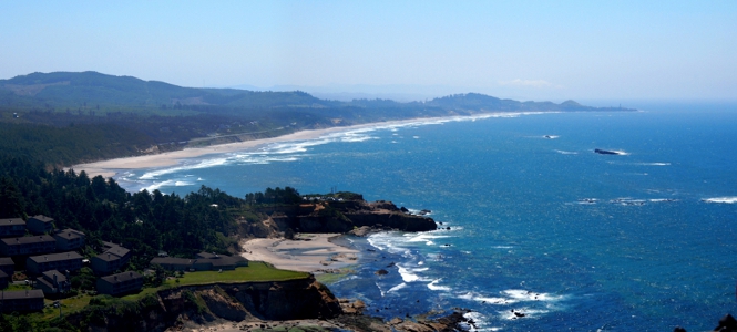 [Several images stitched together to show the view from nearby cliff of the multi-pronged landforms creating the bay on the sunny day. This low tide view makes and sand rocks in the water visible.]